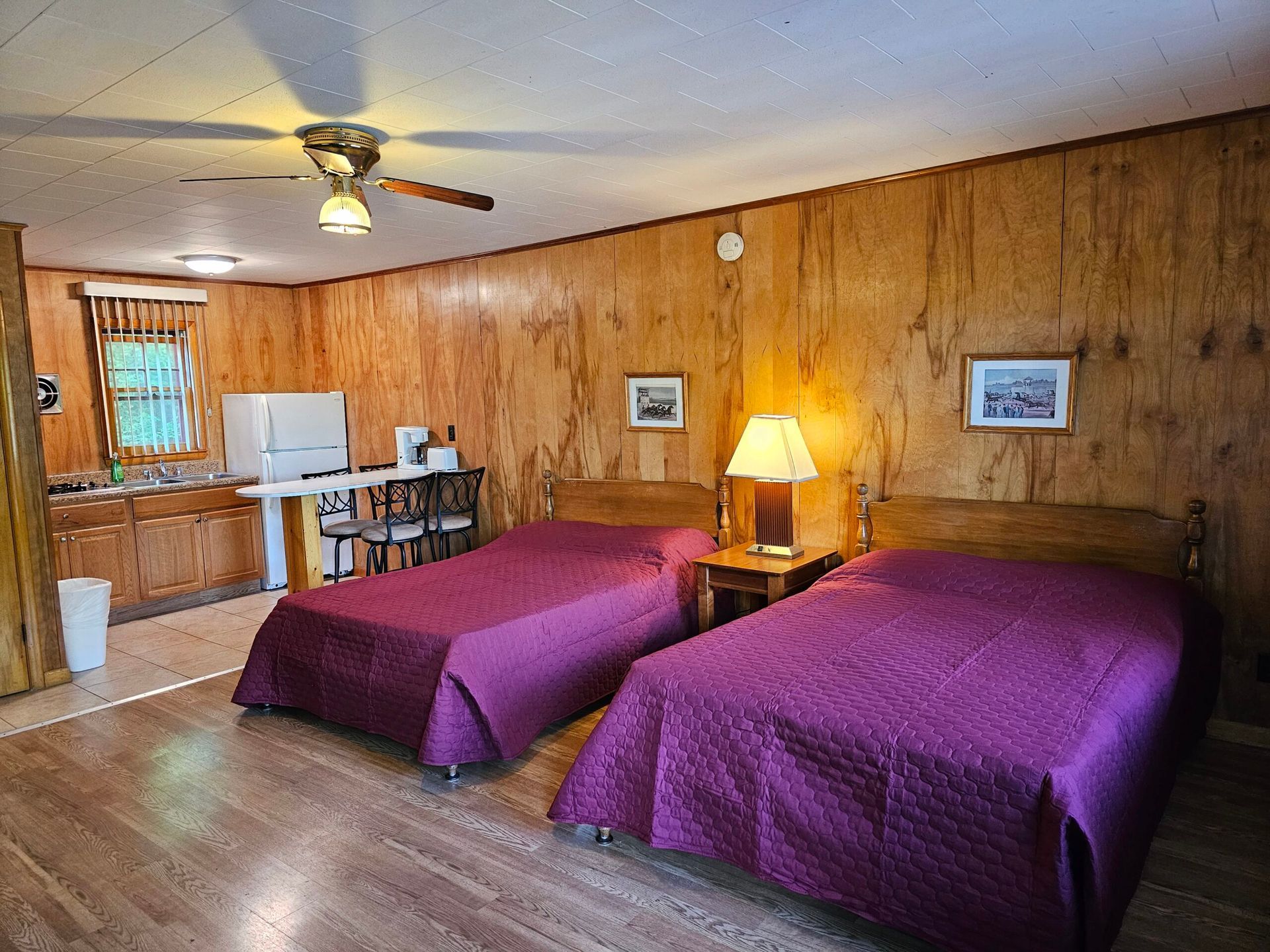 Wood-paneled motel room with two purple-covered beds, a lamp, and a small table by the window.