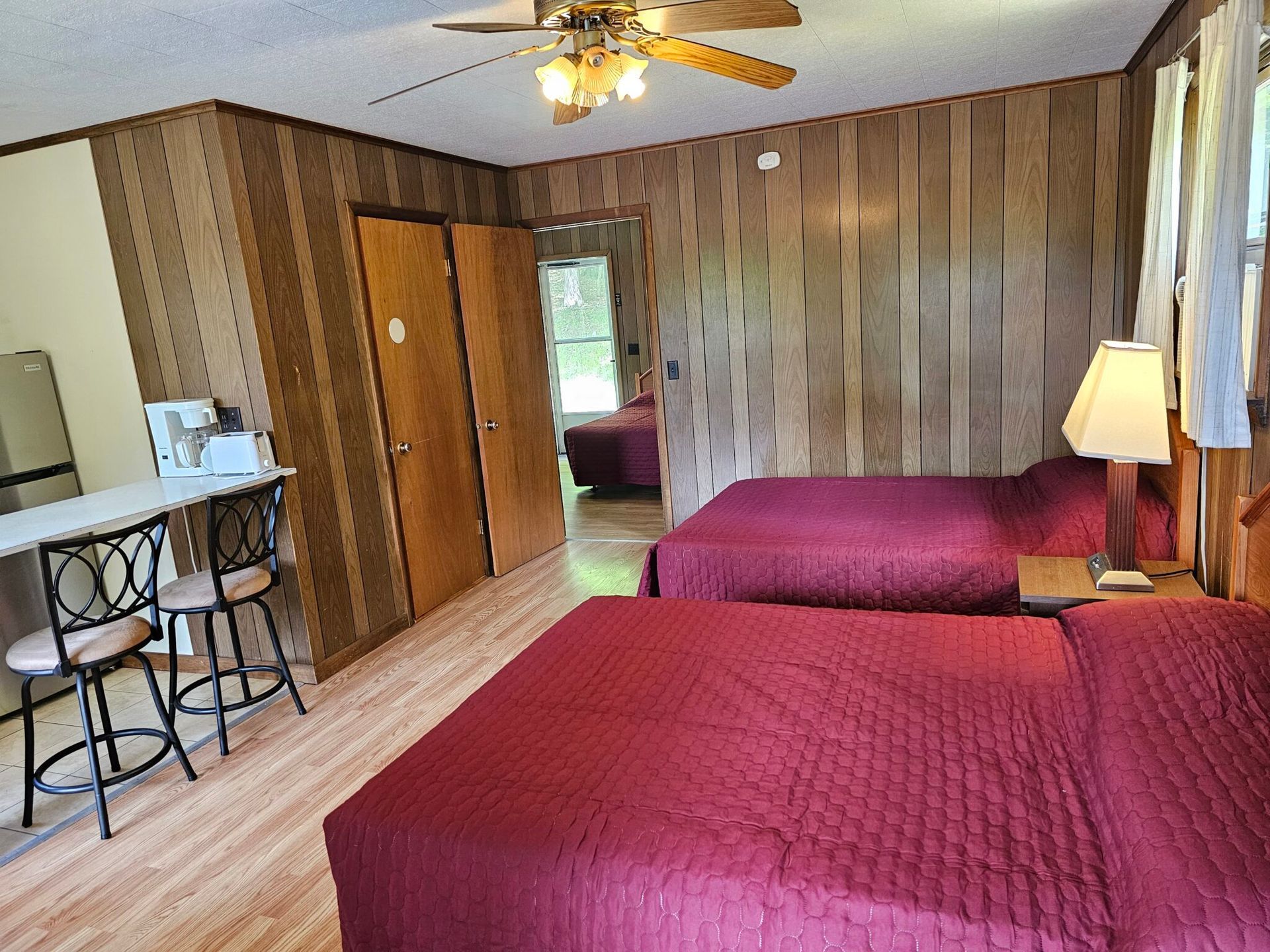 Motel room with two red beds, wood-paneled walls, ceiling fan, lamp, and small table with stools.
