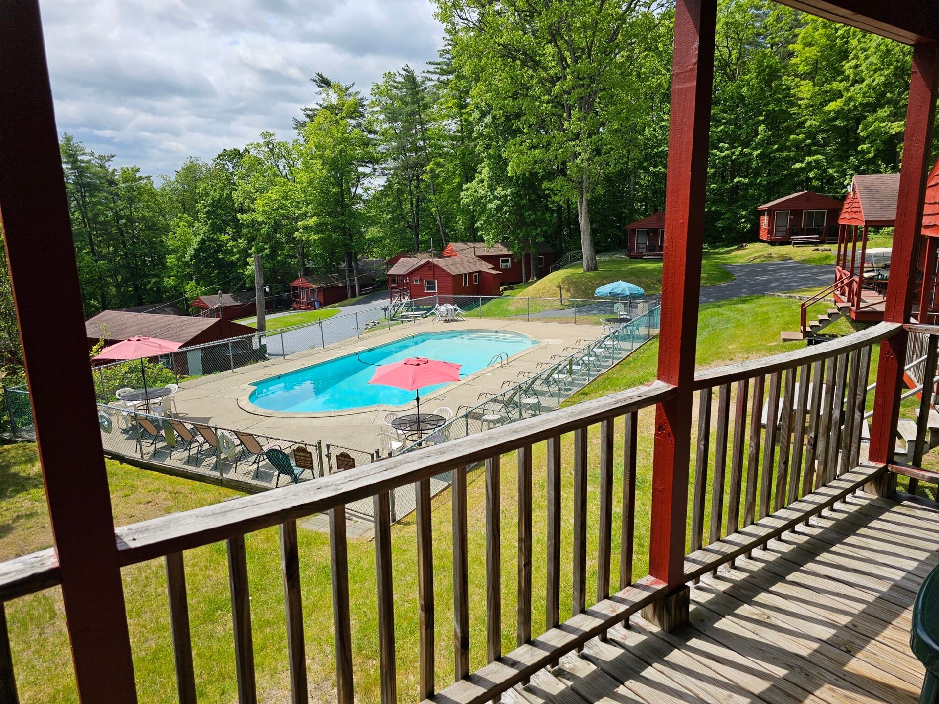 Resort pool with pink umbrellas, seen from a shaded wooden balcony surrounded by trees