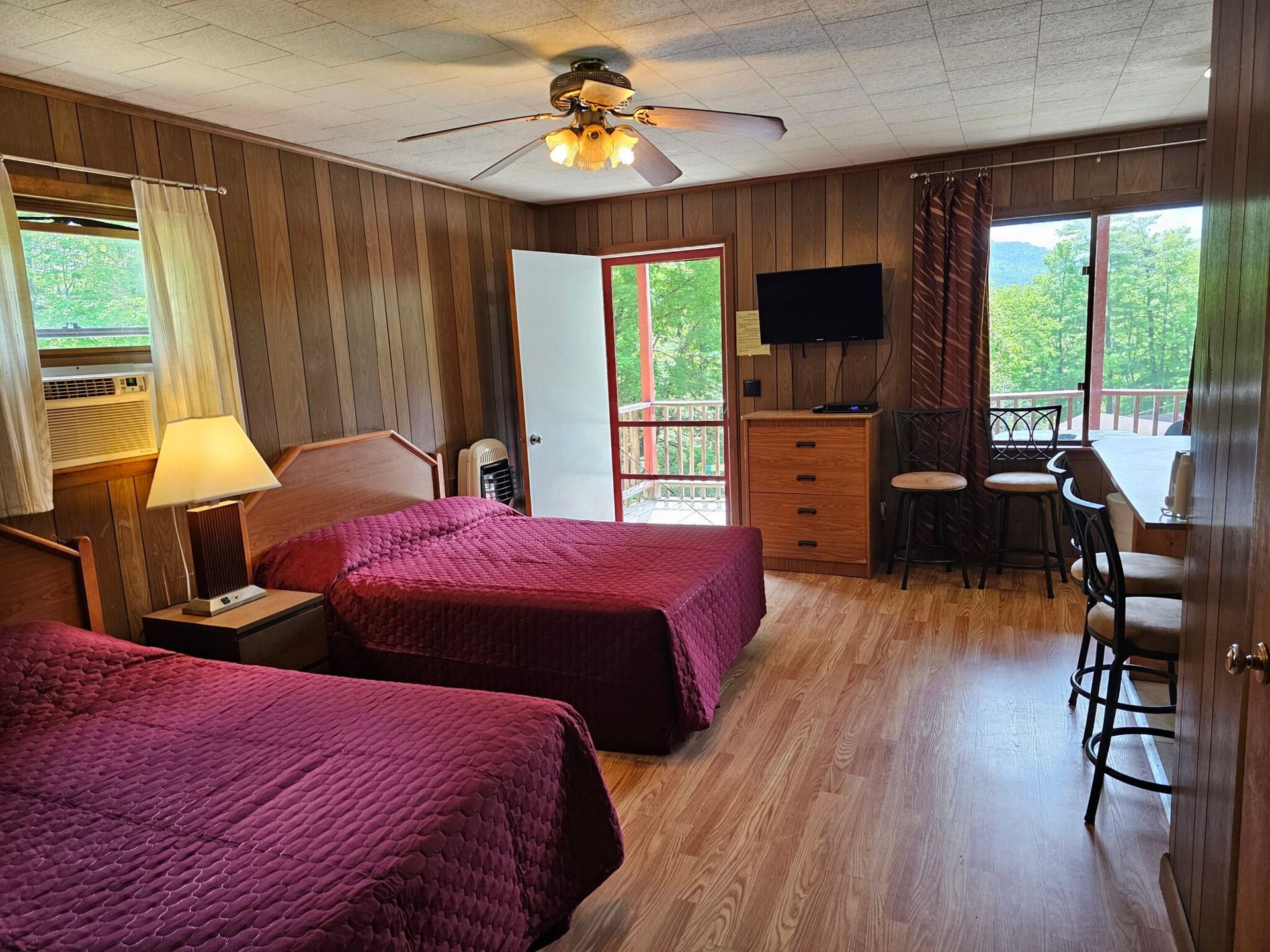 Motel room with two beds, wooden walls, TV, desk, and balcony door with forest view.