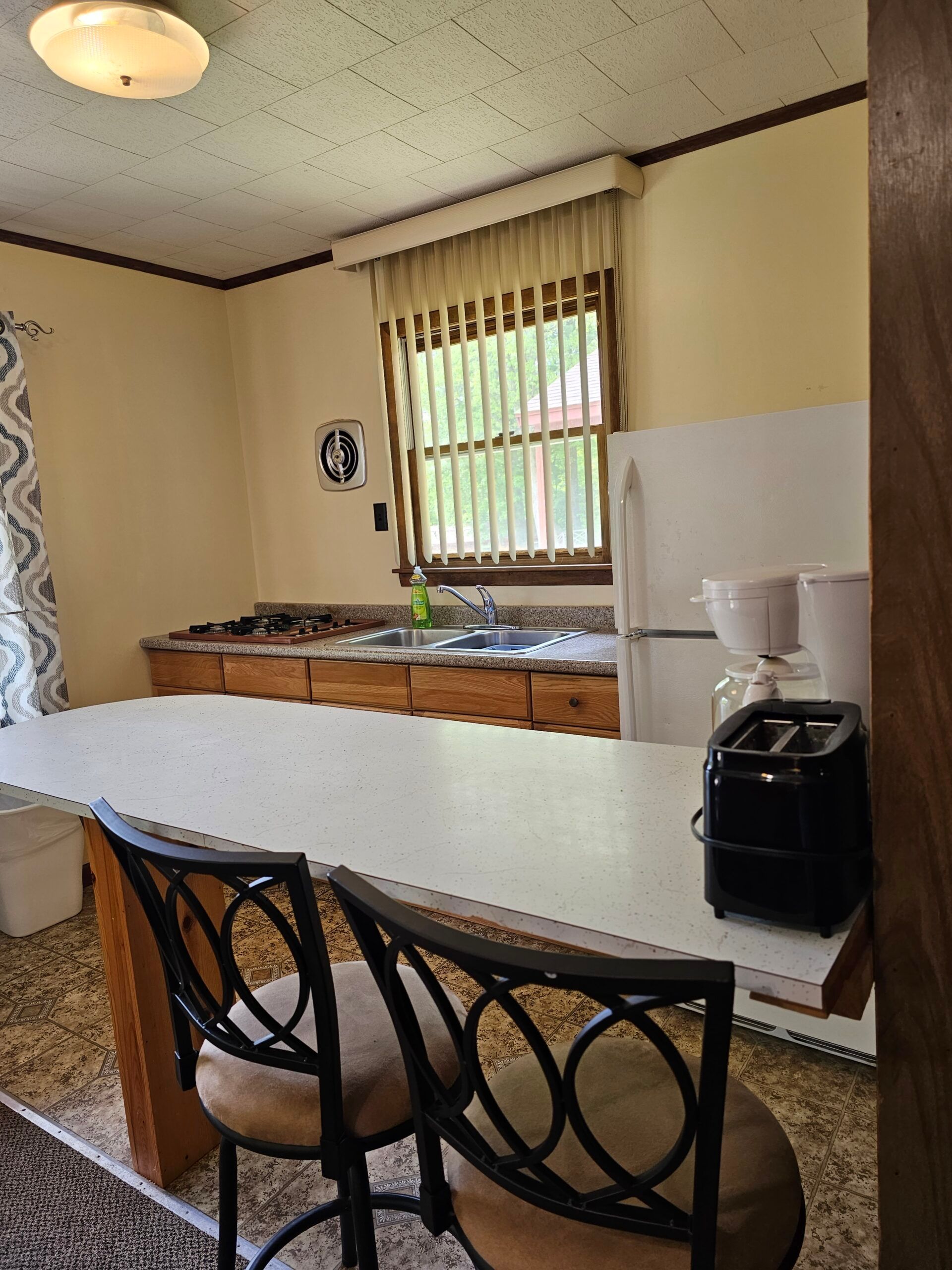 Small kitchen with white counter, wooden cabinets, window blinds, and two stools in the foreground.