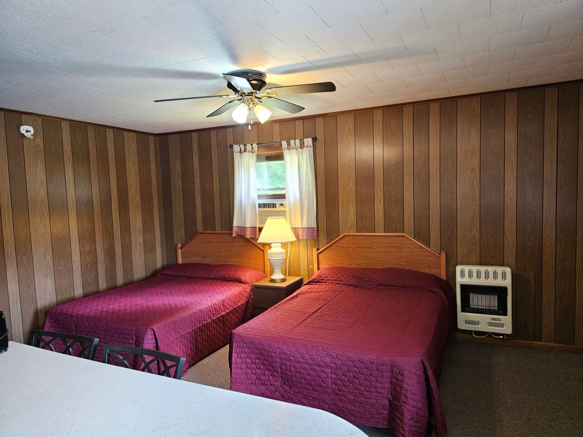 Two twin beds with purple quilts in a wood-paneled motel room, with a lamp and ceiling fan.