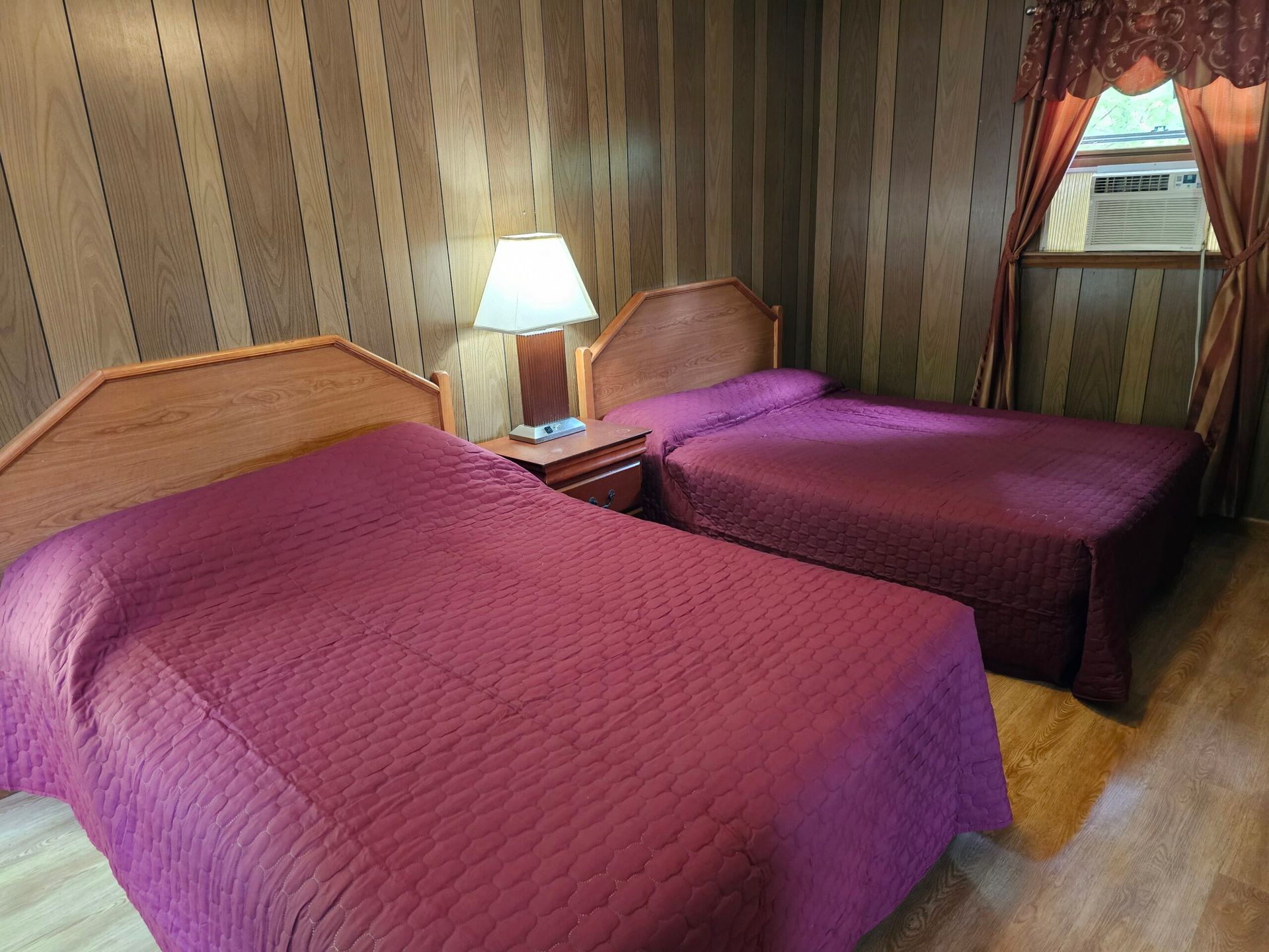Hotel room with two beds covered in purple quilts, a bedside lamp, and a window with curtains.