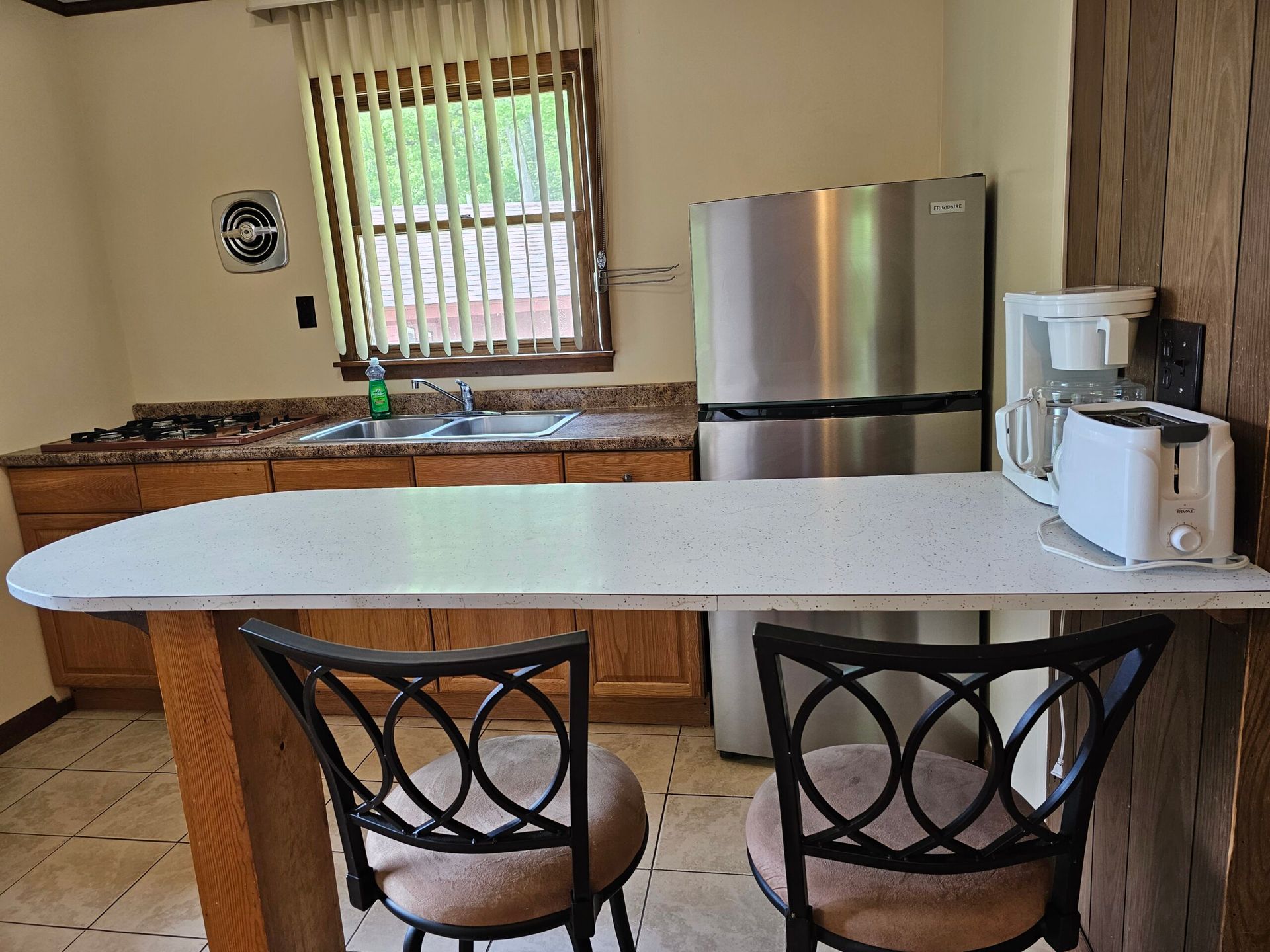Small kitchen with granite island, two bar stools, stainless fridge, coffee maker, and window over the sink.
