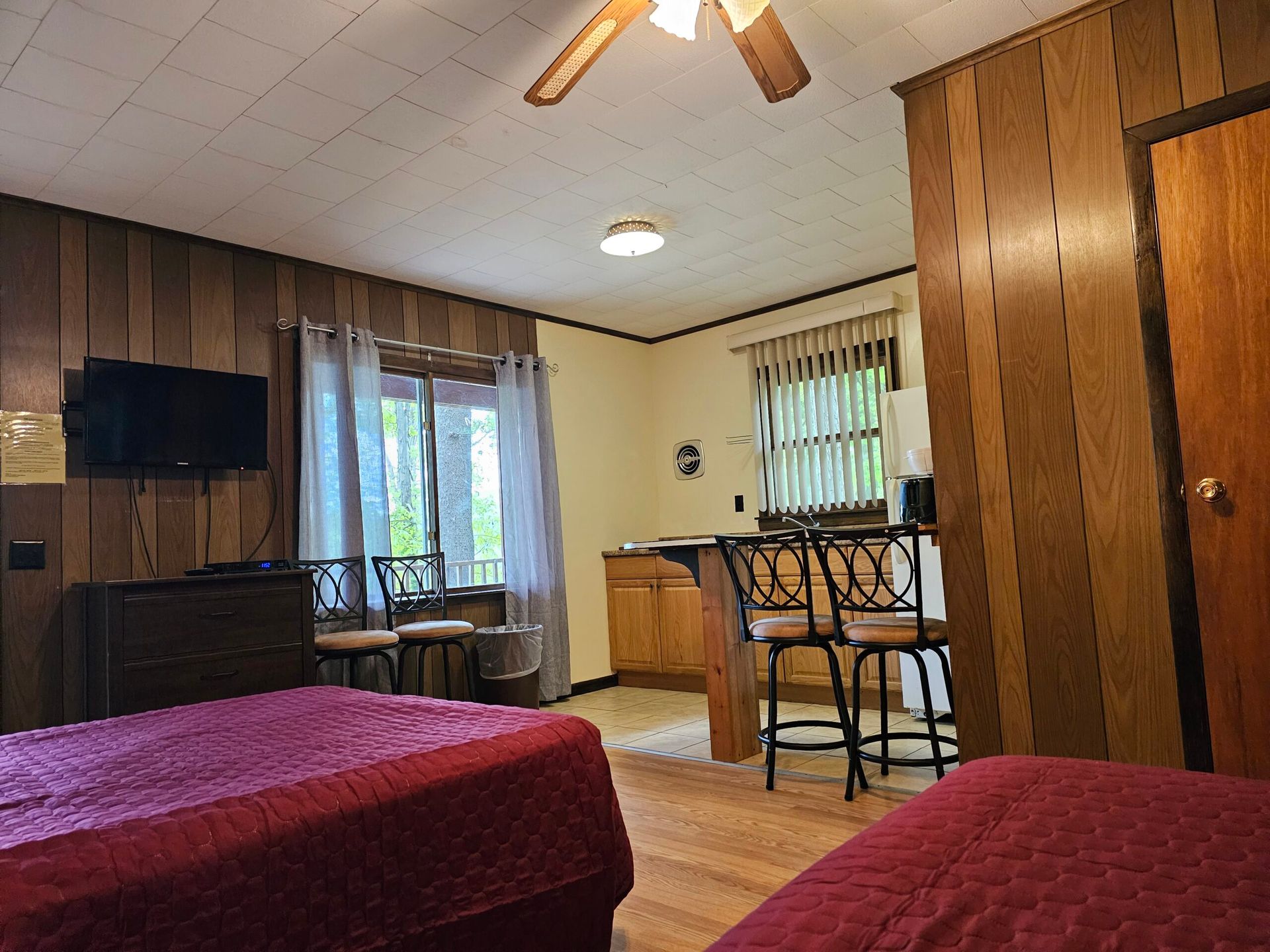 Bedroom with two red-covered beds, wood-paneled walls, TV, ceiling fan, and a small table by the window
