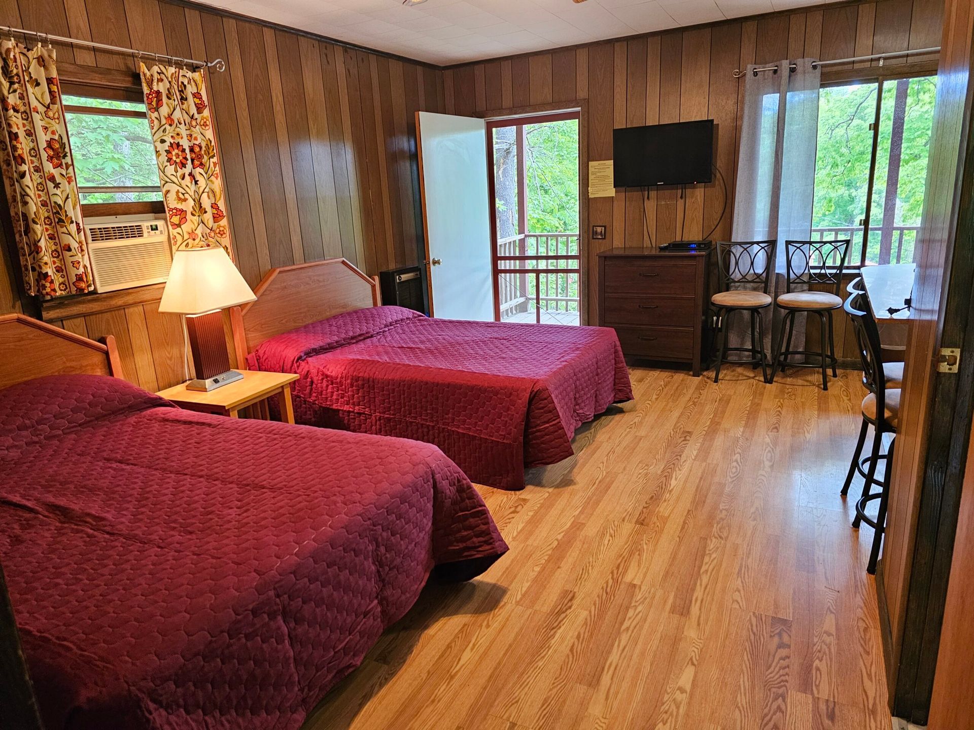 Lodge room with two red beds, wood-paneled walls, a lamp, desk, and balcony door with forest view