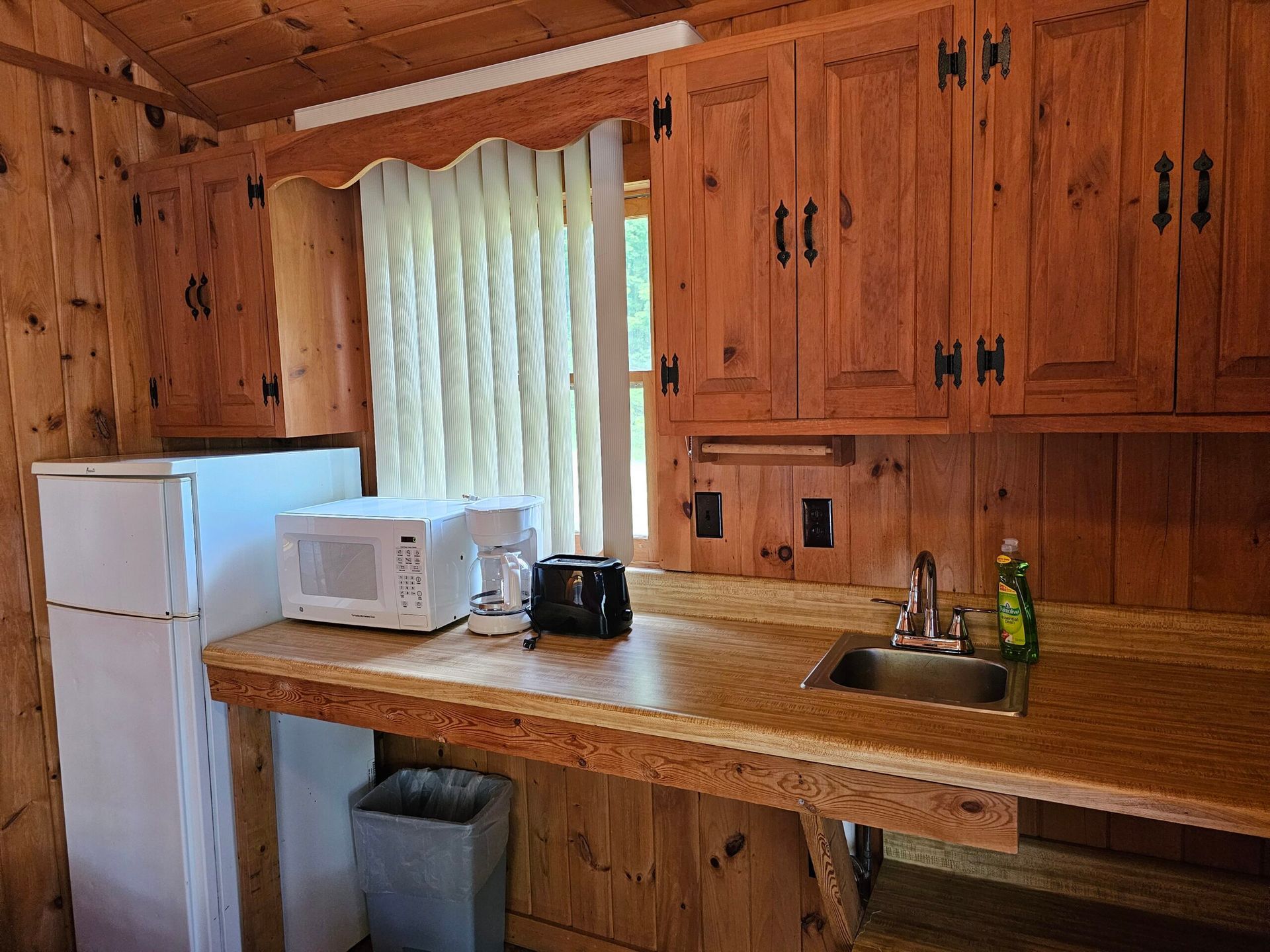 Small wooden kitchen with fridge, microwave, coffee maker, sink, and window curtain