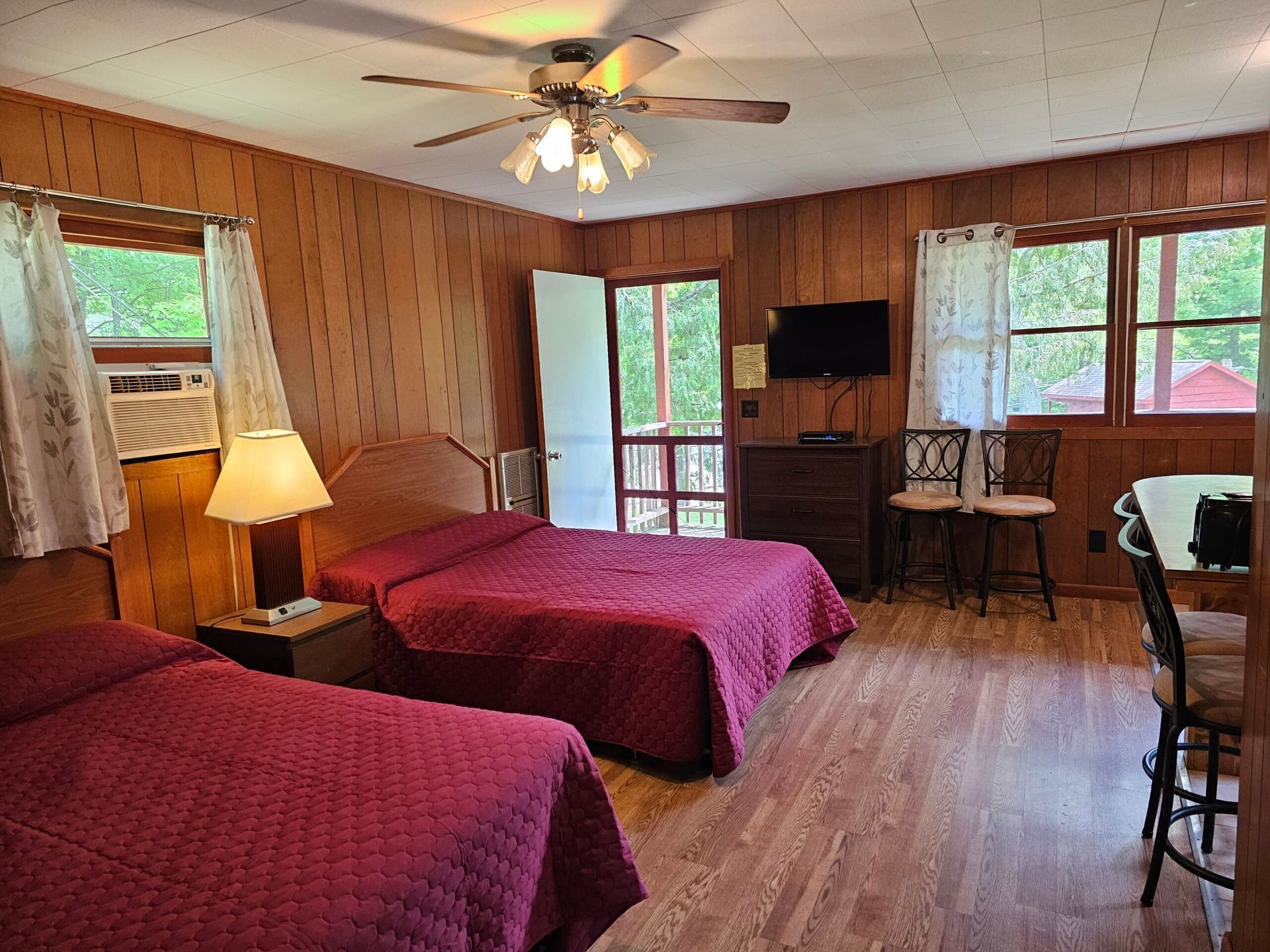 Wood-paneled motel room with two red beds, TV, ceiling fan, and windows overlooking greenery