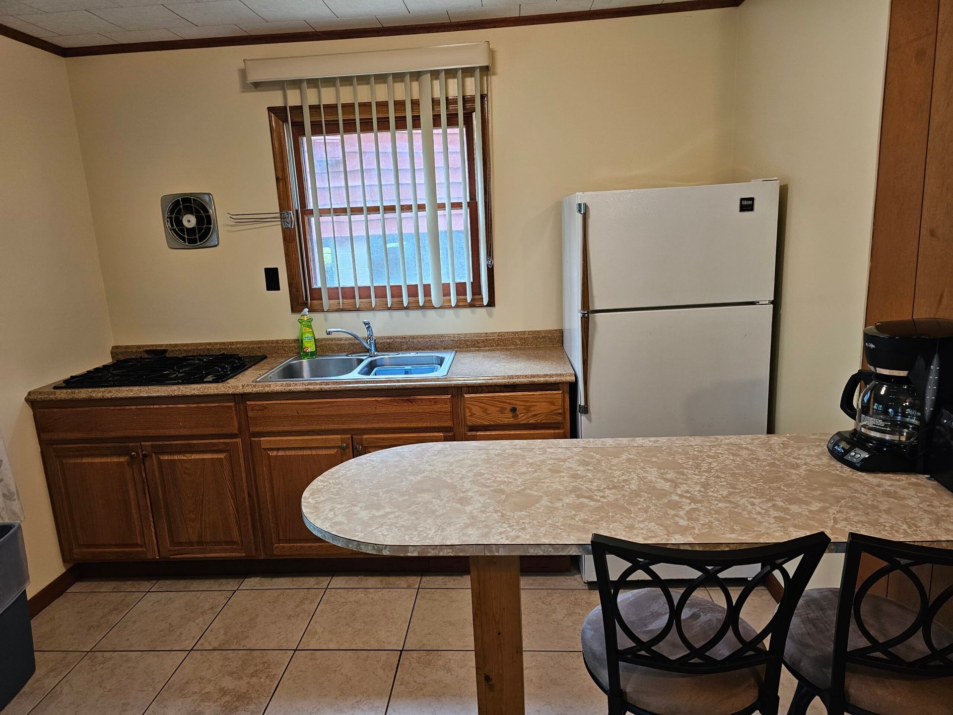 Small kitchen with wooden cabinets, tiled floor, white fridge, and a speckled island table with chairs.