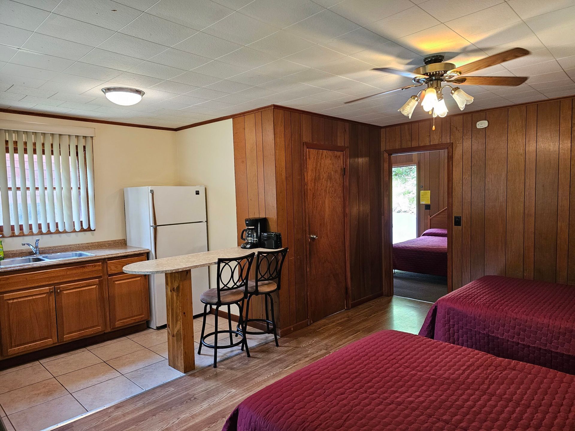 Studio apartment with wood-paneled walls, kitchen, twin beds, and a ceiling fan