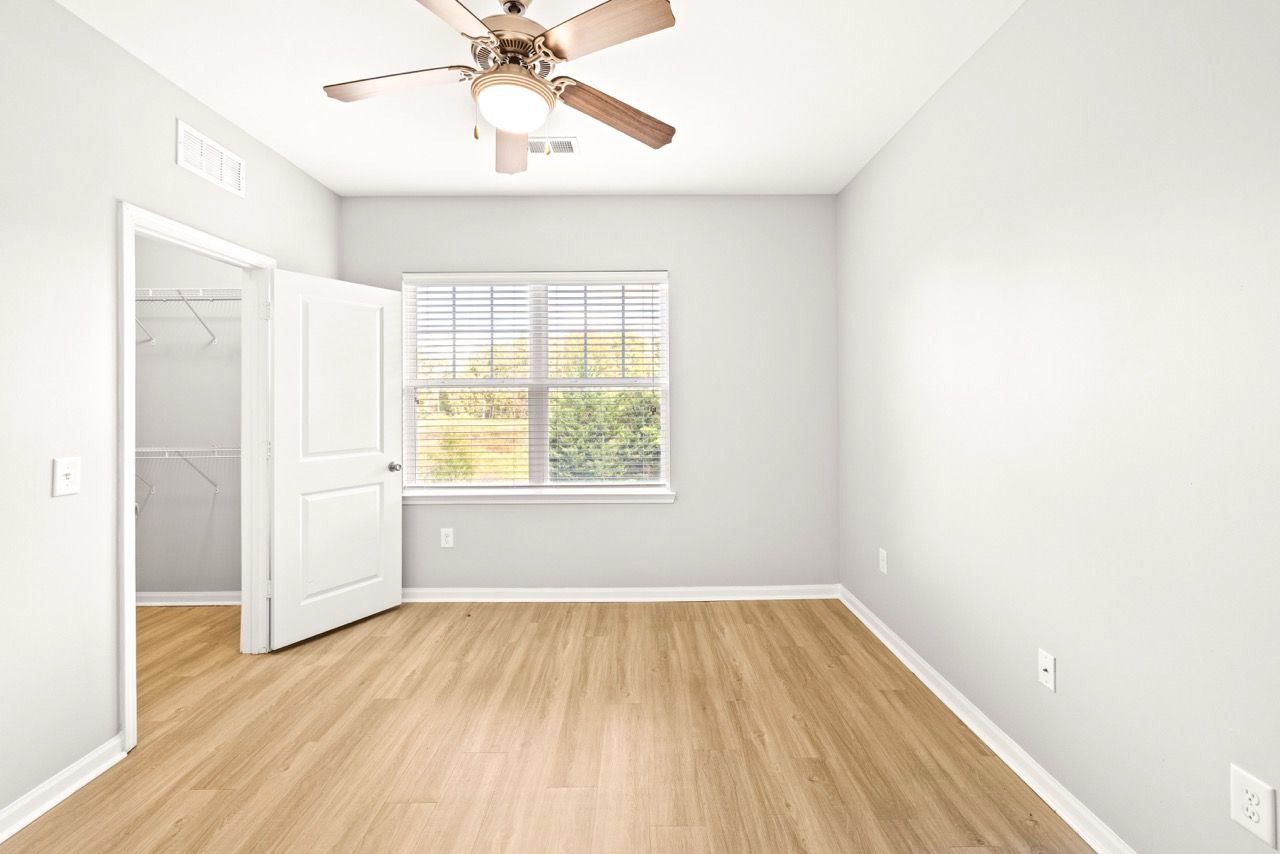 Empty bedroom with wood floors, a window with blinds, ceiling fan, and open closet.