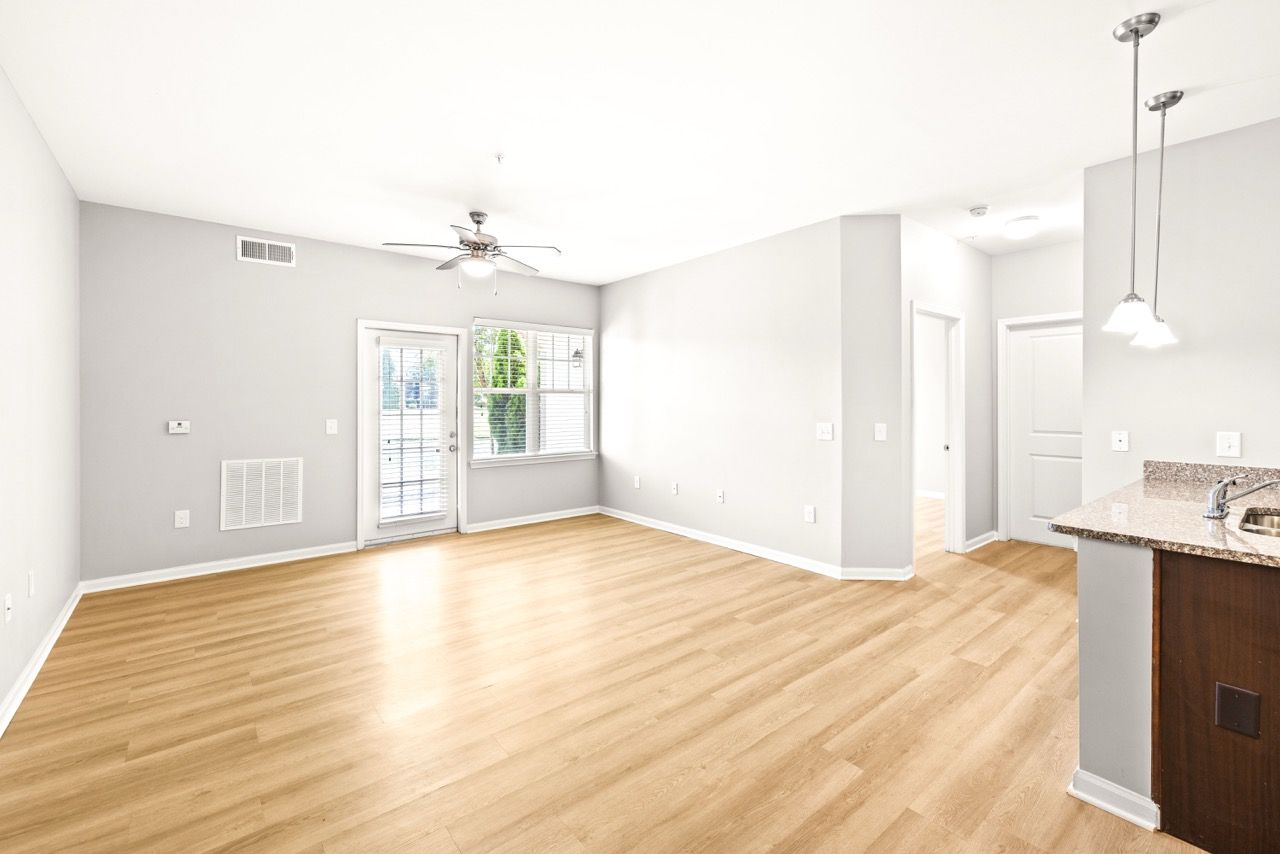 Bright open living room with light gray walls, wood-like flooring, ceiling fan, and kitchen peninsula.