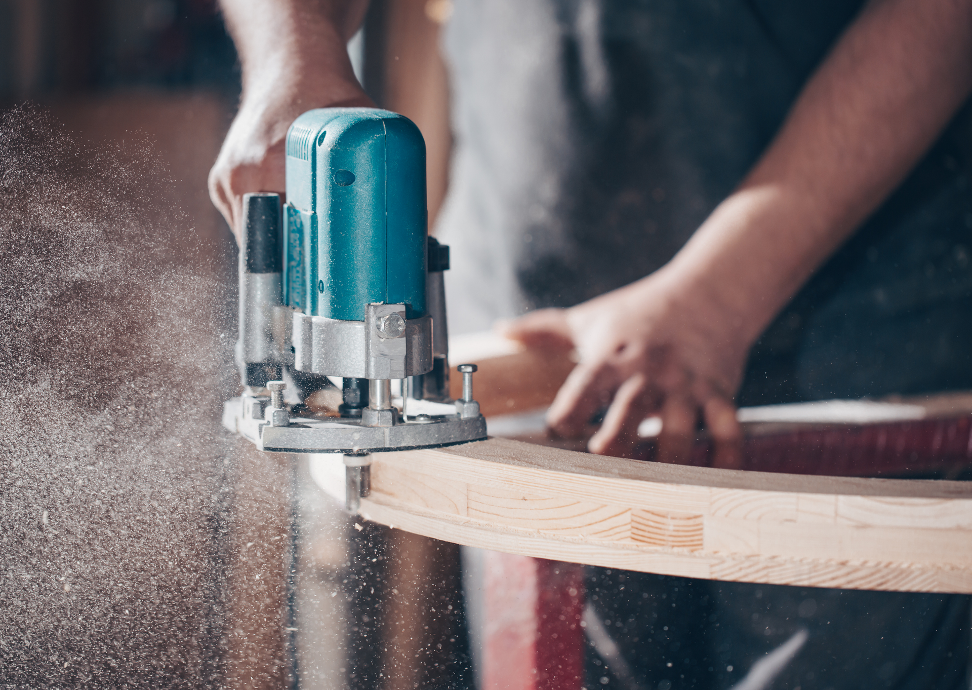Carpenter using a turquoise router on a curved wooden piece, creating sawdust.