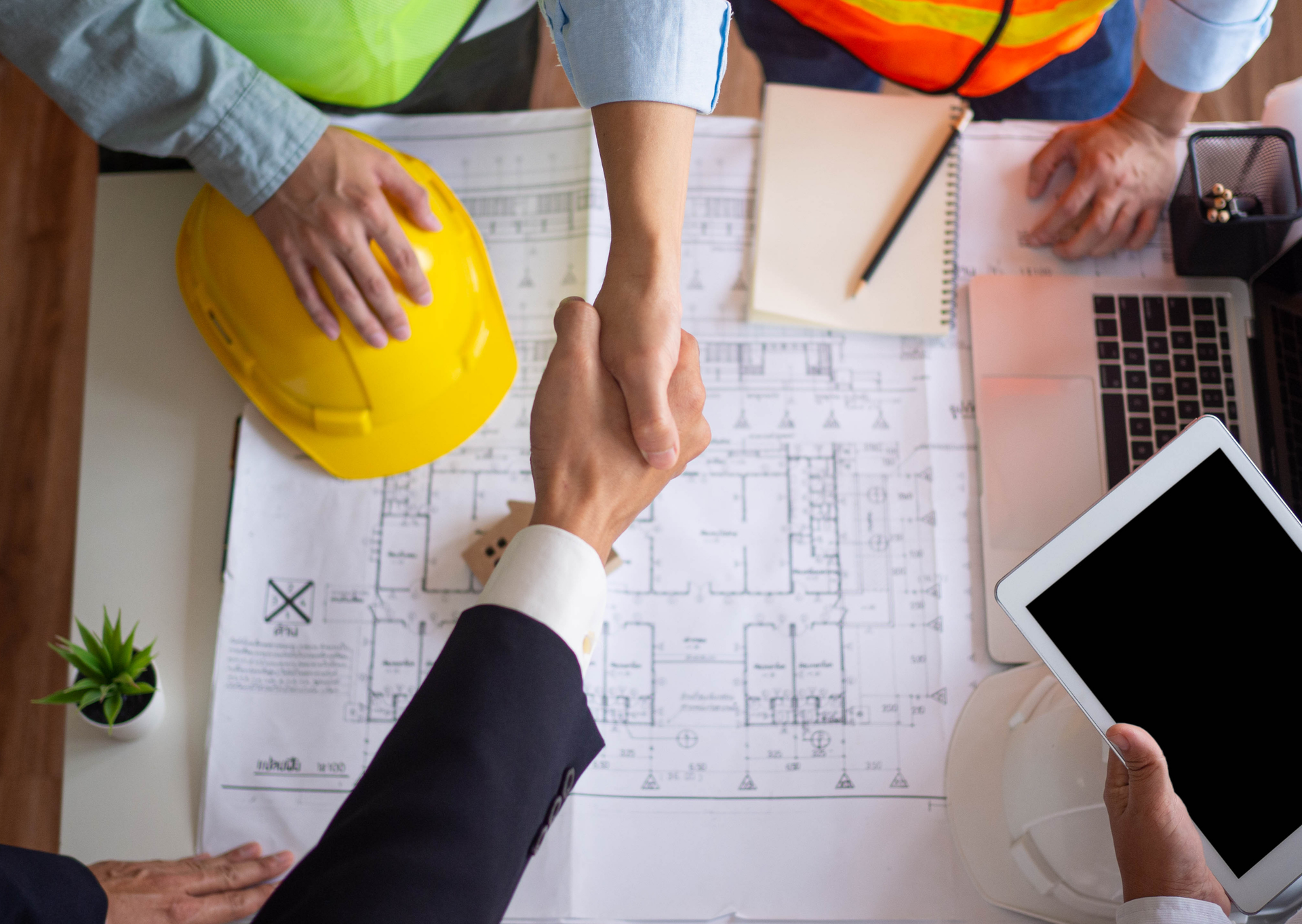 Two people shaking hands over building plans; construction site, agreement.