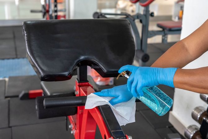 Staff using wet wipe and disinfectant from the bottle spraying sit up bench in gym.