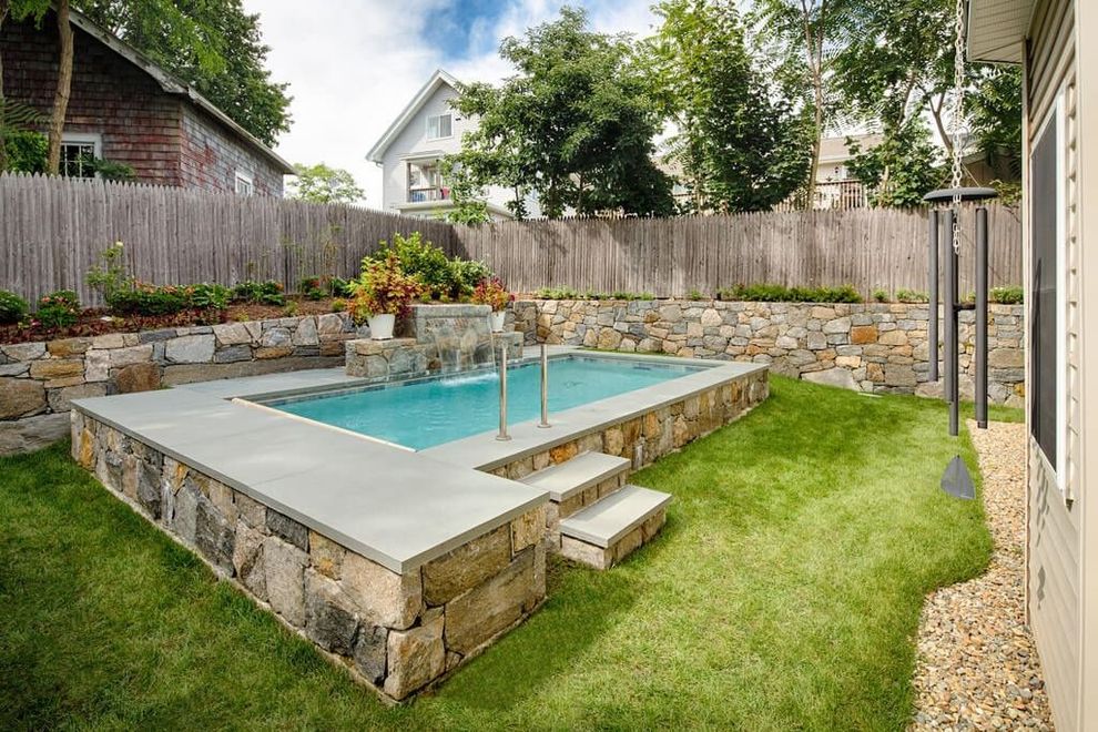 An elevated stone-clad rectangular swimming pool sits in a residential backyard with a grassy lawn and stone walls.