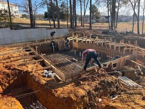 Workers prepare a wooden foundation form and rebar grid at an outdoor construction site with excavated soil trenches.