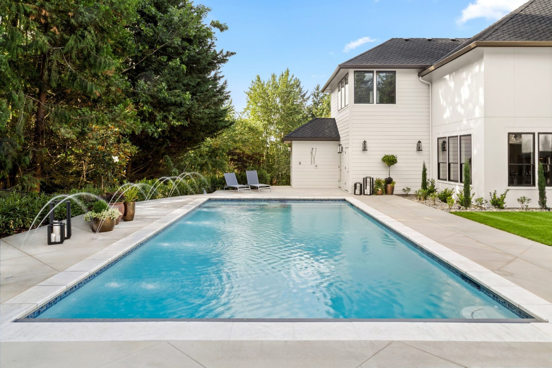 A backyard swimming pool with fountains along the side, adjacent to a white two-story house and a manicured lawn.
