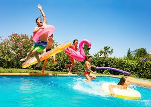 A group of children play in a sunlit swimming pool with colorful inflatable pool toys, jumping and splashing.