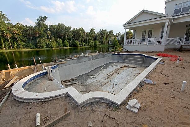 A half-constructed swimming pool with white coping sits in a dirt backyard next to a house near a lake.