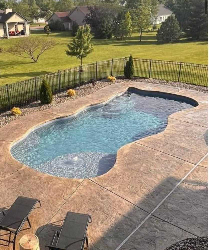 An irregularly shaped blue swimming pool with a textured patio, lounge chairs, and a fenced backyard.