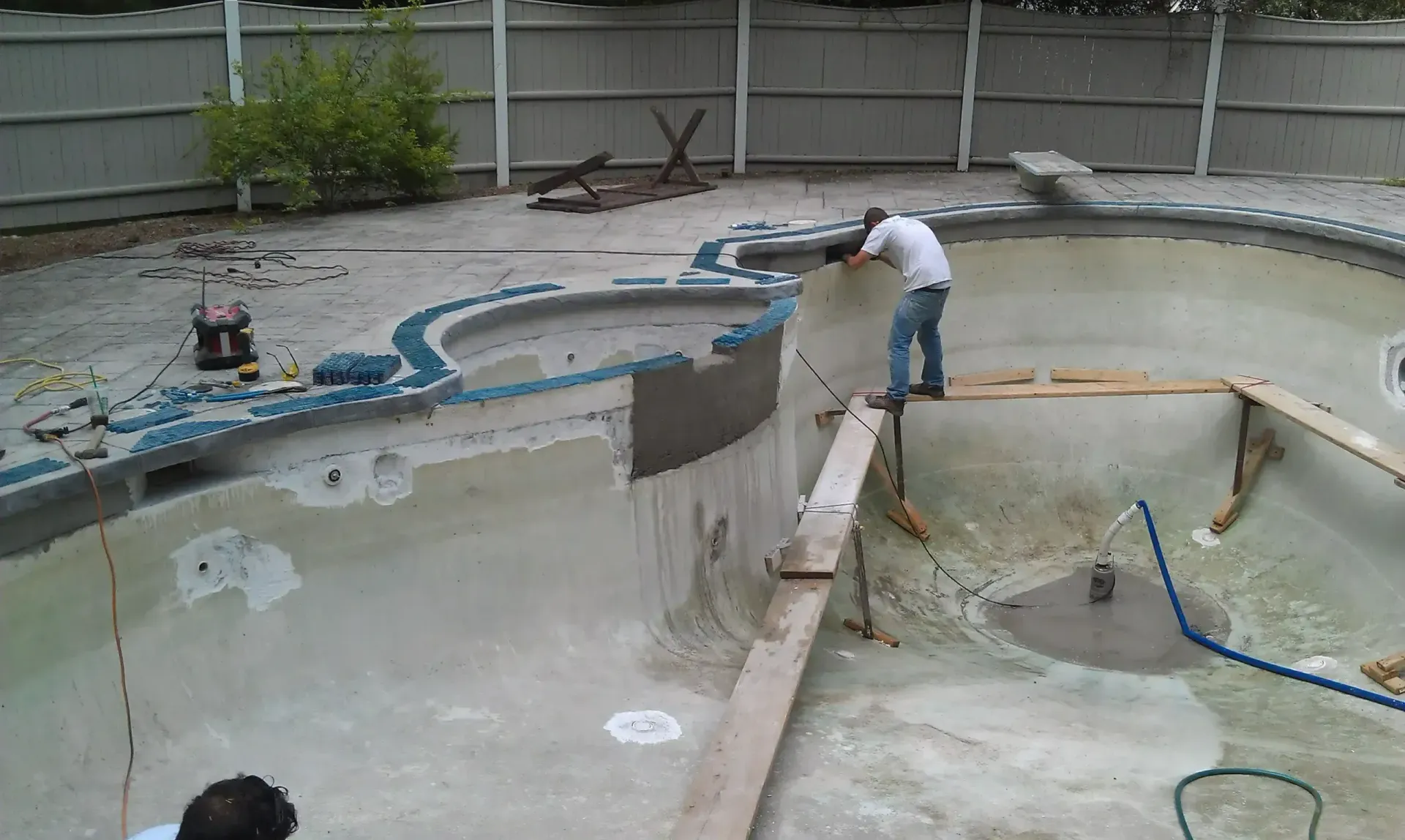 A worker repairs the tile edge of an empty, unfinished swimming pool inside a fenced backyard.