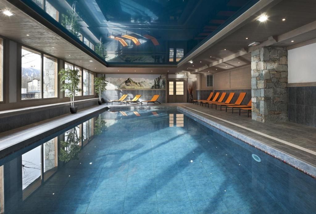 A sleek indoor swimming pool with blue water, floor-to-ceiling windows, and orange lounge chairs on a stone deck.