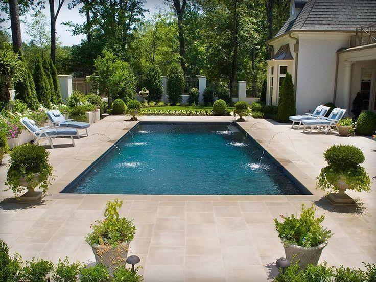 A rectangular swimming pool with fountains sits on a stone patio beside a house, surrounded by lush trees and planters.