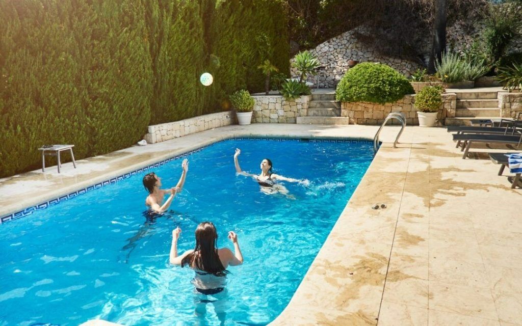 Three people play with a white ball in a sunny outdoor swimming pool surrounded by greenery and stone tiling.