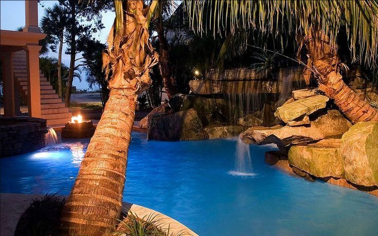 A backyard swimming pool at dusk with illuminated waterfalls, rock landscaping, and palm trees.