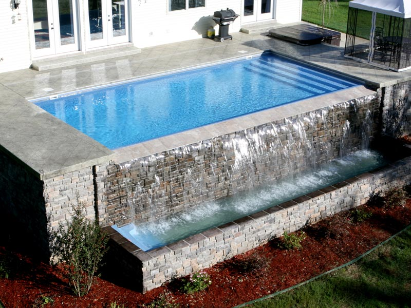 A backyard swimming pool with a stone wall water feature cascading into a lower basin, set near a house patio.