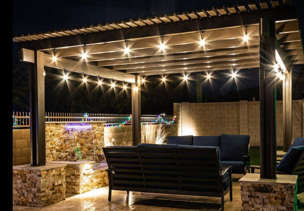 A modern outdoor patio at night featuring a dark wood pergola illuminated by string lights over blue cushioned seating.