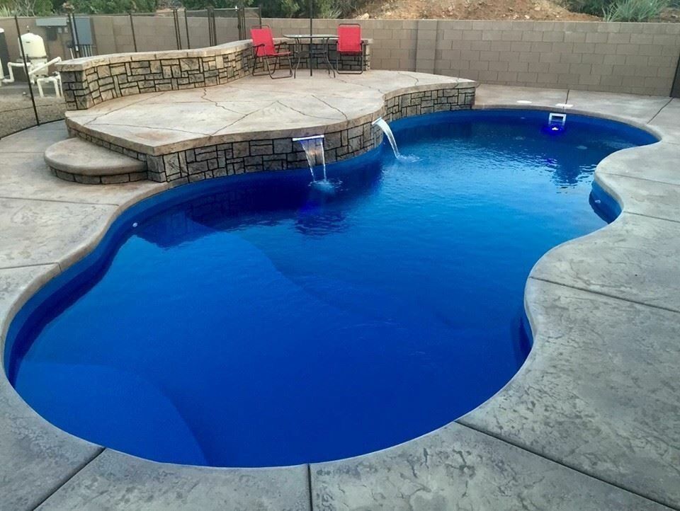 An oval swimming pool with deep blue water, stone-tiered patio seating, and two cascading water features in a backyard.