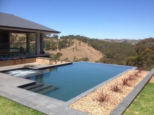 An infinity pool overlooking a sunlit valley, adjacent to a modern home with a covered patio and stone accents.