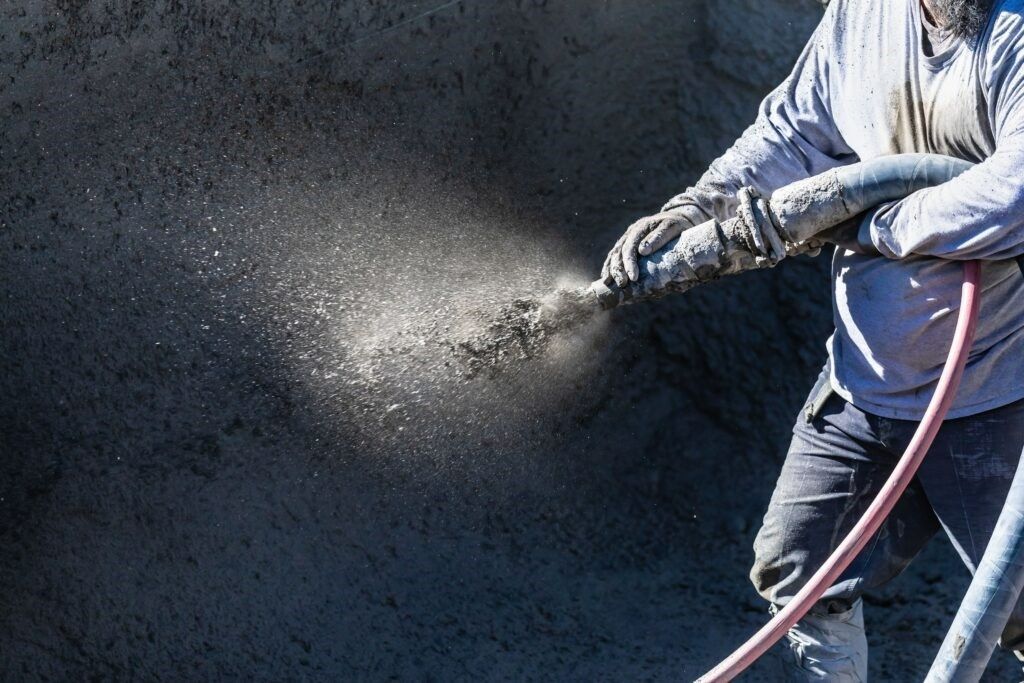 A worker sprays concrete onto a surface using a high-pressure nozzle and hose.