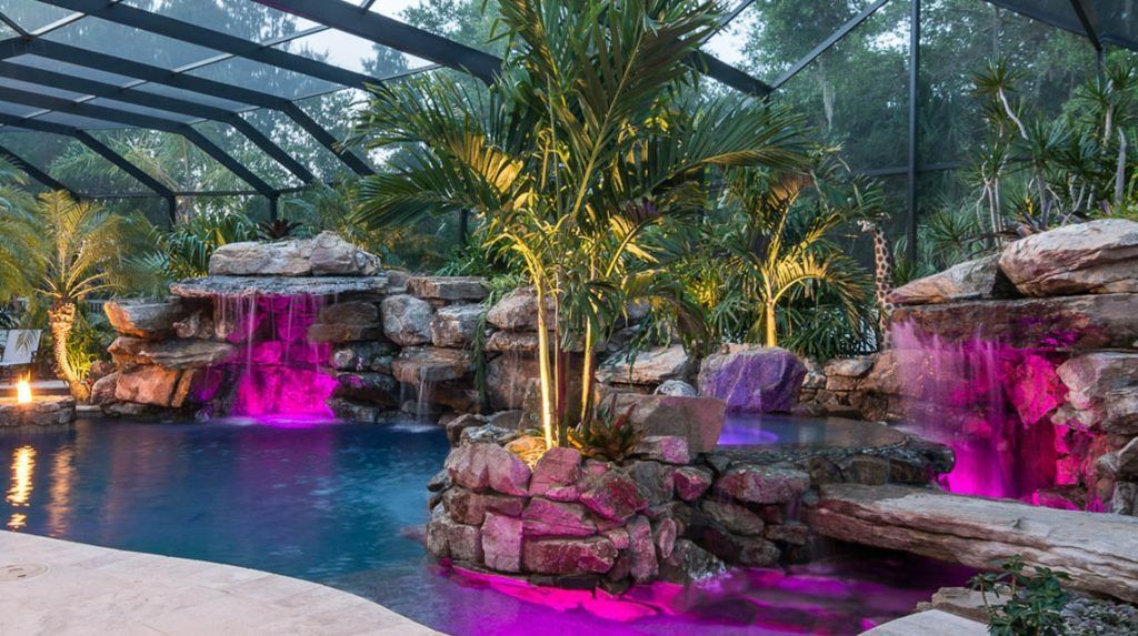 A luxury swimming pool with a rock waterfall feature, palm trees, and vibrant magenta underwater lighting.