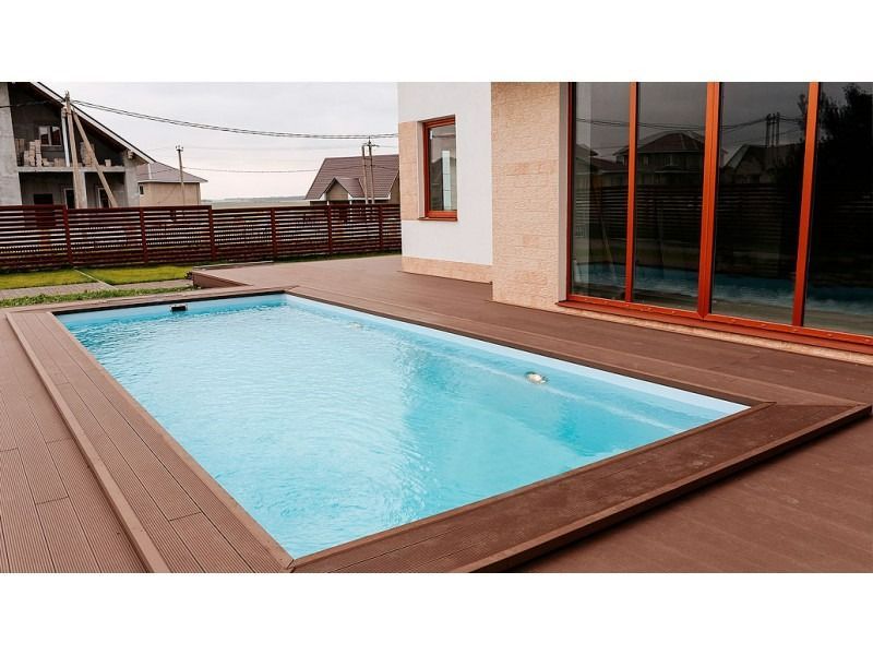A rectangular swimming pool with light blue water sits on a brown composite deck next to a modern house.