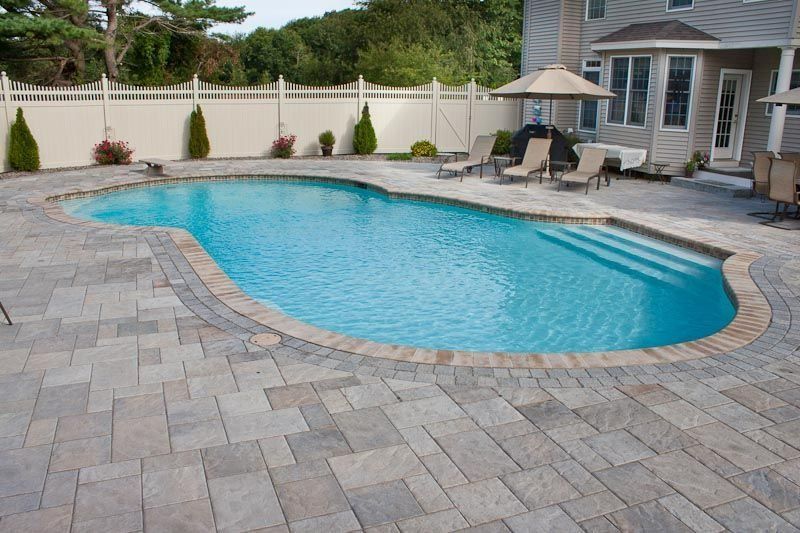 A backyard swimming pool with a stone paver patio, lounge chairs, an umbrella, and a tall fence on a sunny day.