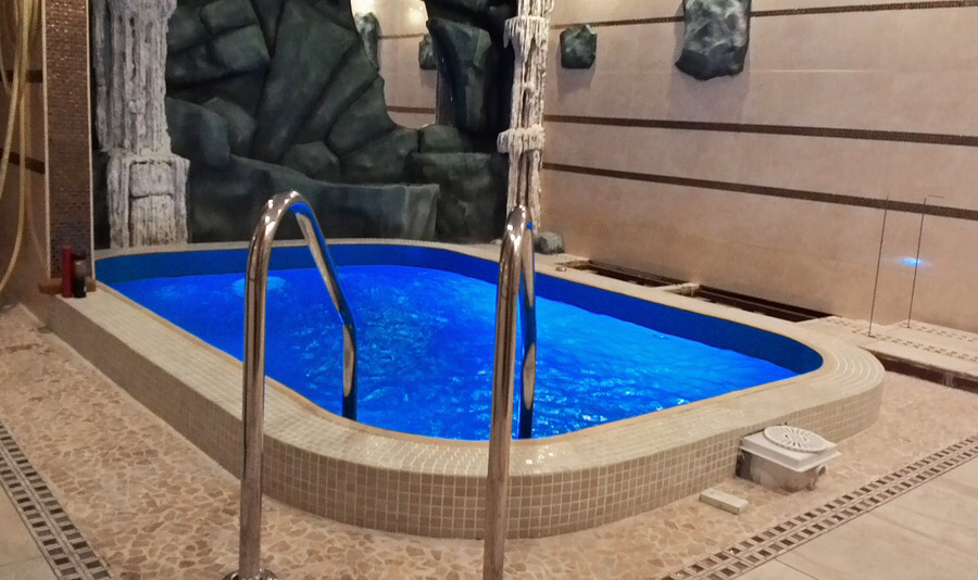 An indoor rectangular swimming pool with bright blue water, surrounded by decorative stone walls and light-colored tile.
