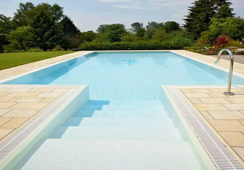 A bright, rectangular swimming pool with light blue water and wide, stepped entry, surrounded by light-colored stone tiles.