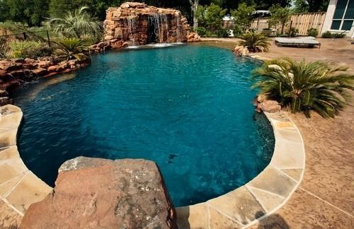 A blue, free-form swimming pool with a rock waterfall feature, surrounded by stone decking and tropical landscaping.