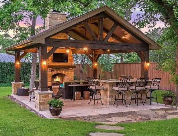A covered outdoor pavilion featuring a stone fireplace, a TV, a bar with stools, and seating in a lush backyard setting.