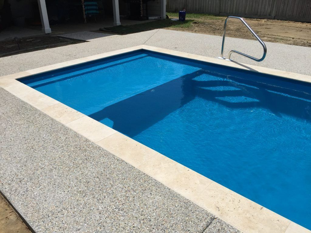 An outdoor rectangular swimming pool with blue water, light stone coping, a speckled concrete deck, and a metal handrail.