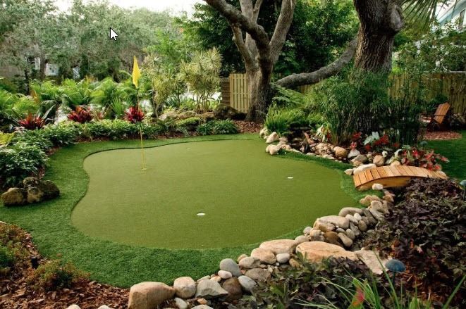 A backyard putting green with a yellow flag, bordered by rocks, lush plants, and a small wooden bridge.