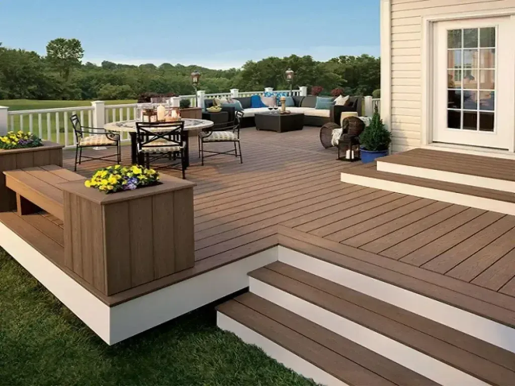A spacious brown composite deck featuring a dining area, cushioned outdoor seating, and built-in planters with flowers.