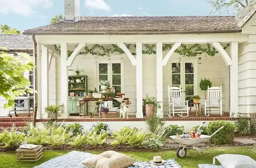 A white porch with rockers and a table decorated with plants, overlooking a lush garden with a picnic blanket in front.