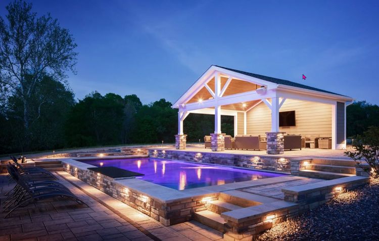 A glowing, rectangular swimming pool with blue light at night, set beside a stone-columned patio with a covered pavilion.