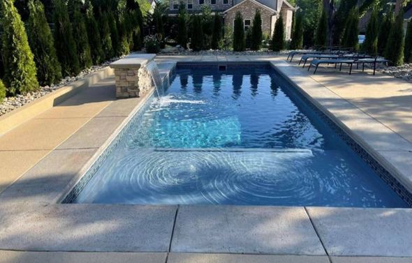 A rectangular backyard swimming pool with a stone water feature, light concrete decking, and evergreen trees in the back.