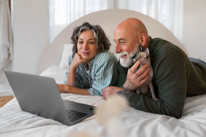 Couple looking at laptop, holding a small dog, on a bed.