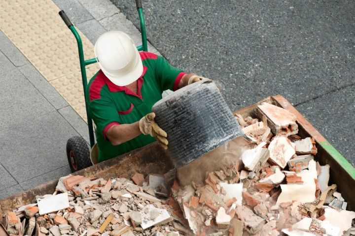 A construction worker wearing a white hard hat and green uniform empties a bucket of rubble into a skip.